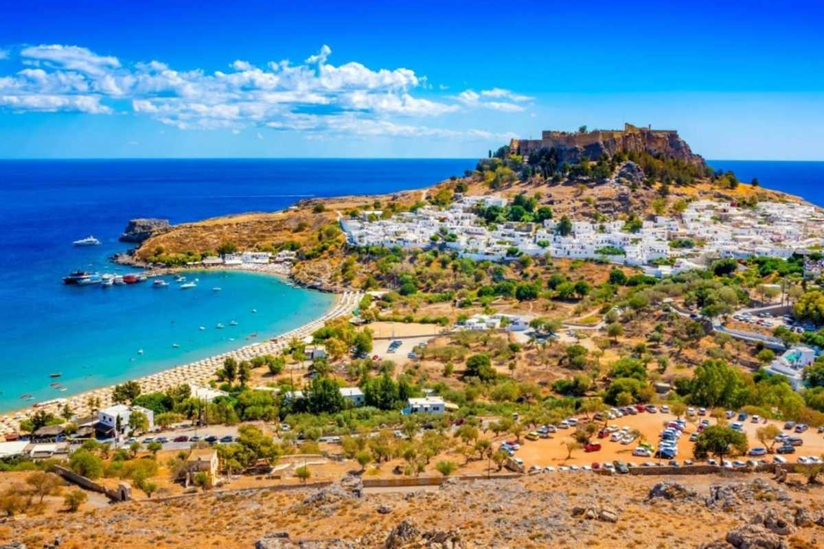 Lindos day trip from Rhodes: the Acropolis of Lindos on its rocky hill above the whitewashed village, with a curved sandy bay and turquoise Aegean Sea on the left