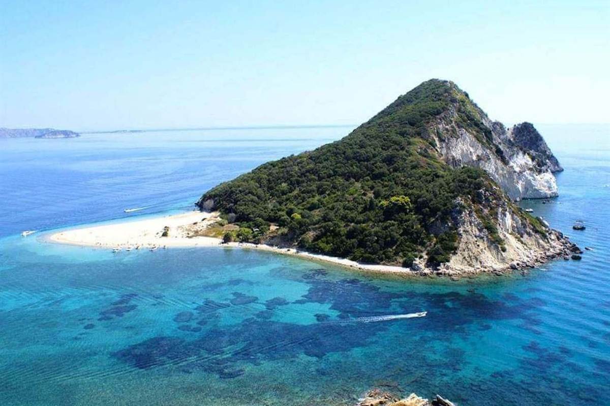 Marathonisi turtle island tour: Marathonisi island seen from above, a green wooded hill with pale limestone cliffs, a curved white sand spit with a few small boats beside it, and clear turquoise and deep blue water of Laganas Bay with one speedboat leaving a white wake