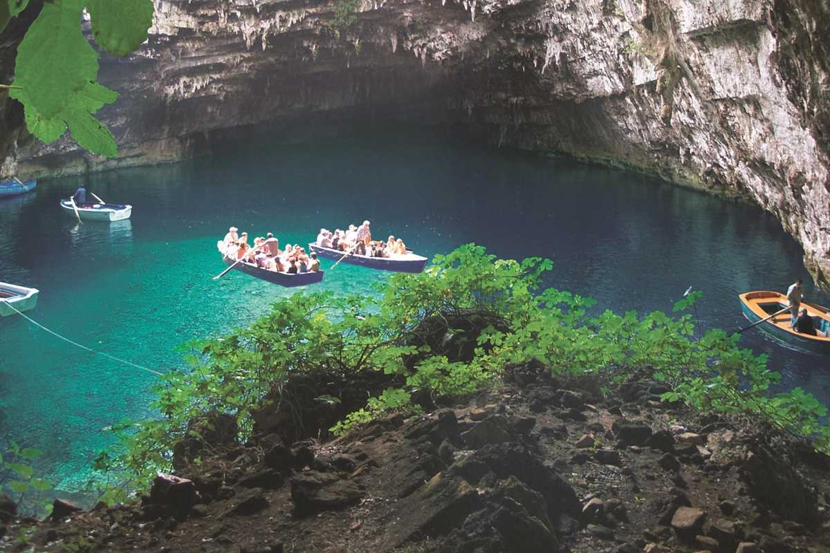 Melissani and Drogarati caves tour: wooden rowing boats carrying visitors across the clear turquoise water of Melissani Lake under a layered limestone cave roof, with green leaves in the foreground and a dark rocky shore below