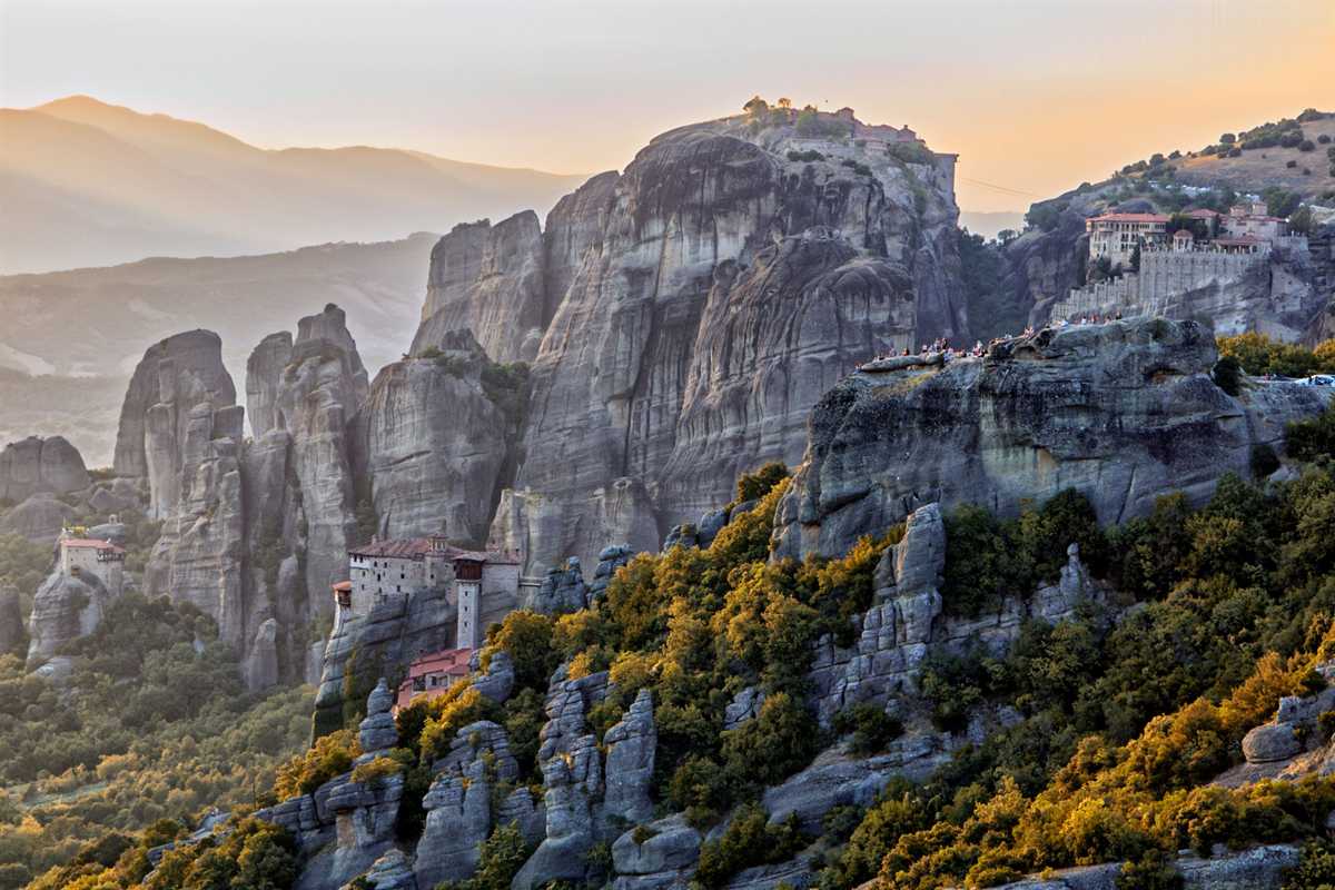 Meteora day trip from Athens: tall grey rock towers in warm evening light with a monastery on top of the large central pillar, a stone monastery with red roofs on a smaller rock at the lower left, more monastery buildings at the upper right and forested slopes in the foreground