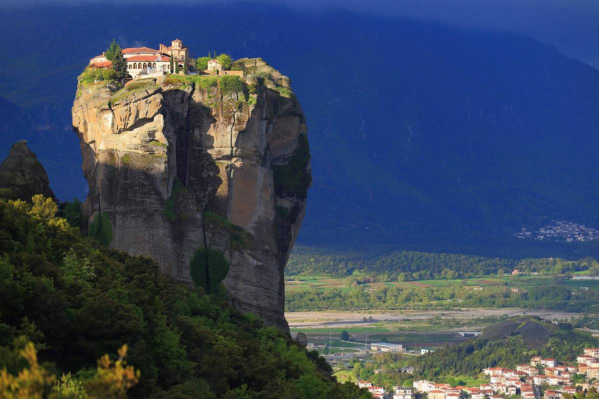 Meteora day trip from Thessaloniki: a monastery with red-tiled roofs and a small bell tower on top of a tall sheer rock pillar, with a dark mountain slope behind, a flat green river valley and the red roofs of a town at the lower right