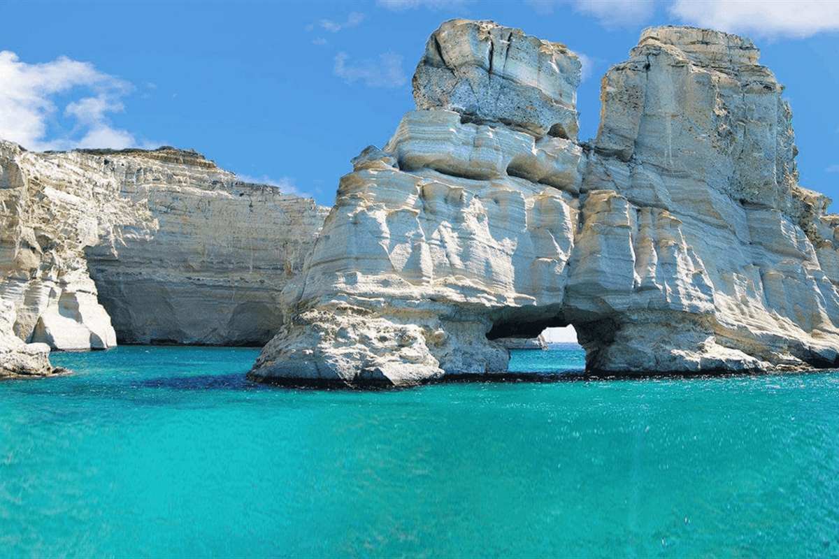Milos private transfer: white volcanic rock cliffs and a tall double rock arch rising from bright turquoise water at Kleftiko under a blue sky with a few white clouds