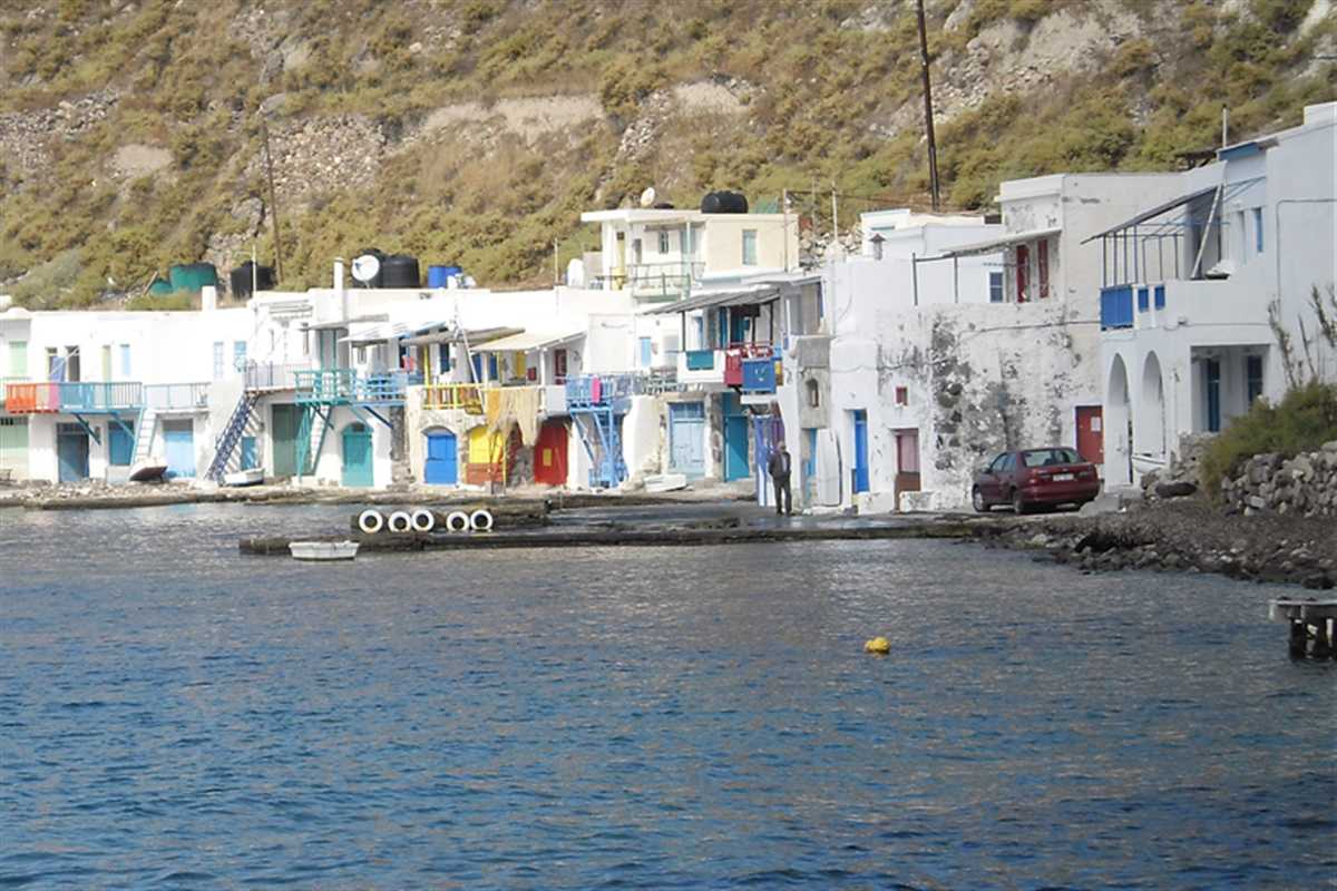 Milos private transfer: a row of white boat-garage houses with blue, red, yellow and green doors and balconies along the water at Klima, below a dry hillside and beside calm blue sea