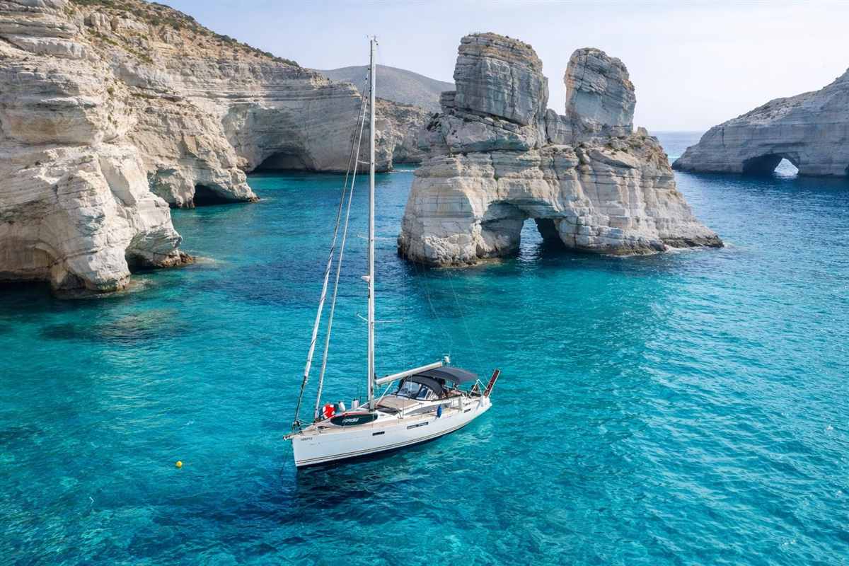 Milos sailing: a white sailboat with one tall mast anchored in bright turquoise water in a bay of Kleftiko, with pale layered rock cliffs, sea caves and a tall natural rock arch behind it under a hazy blue sky