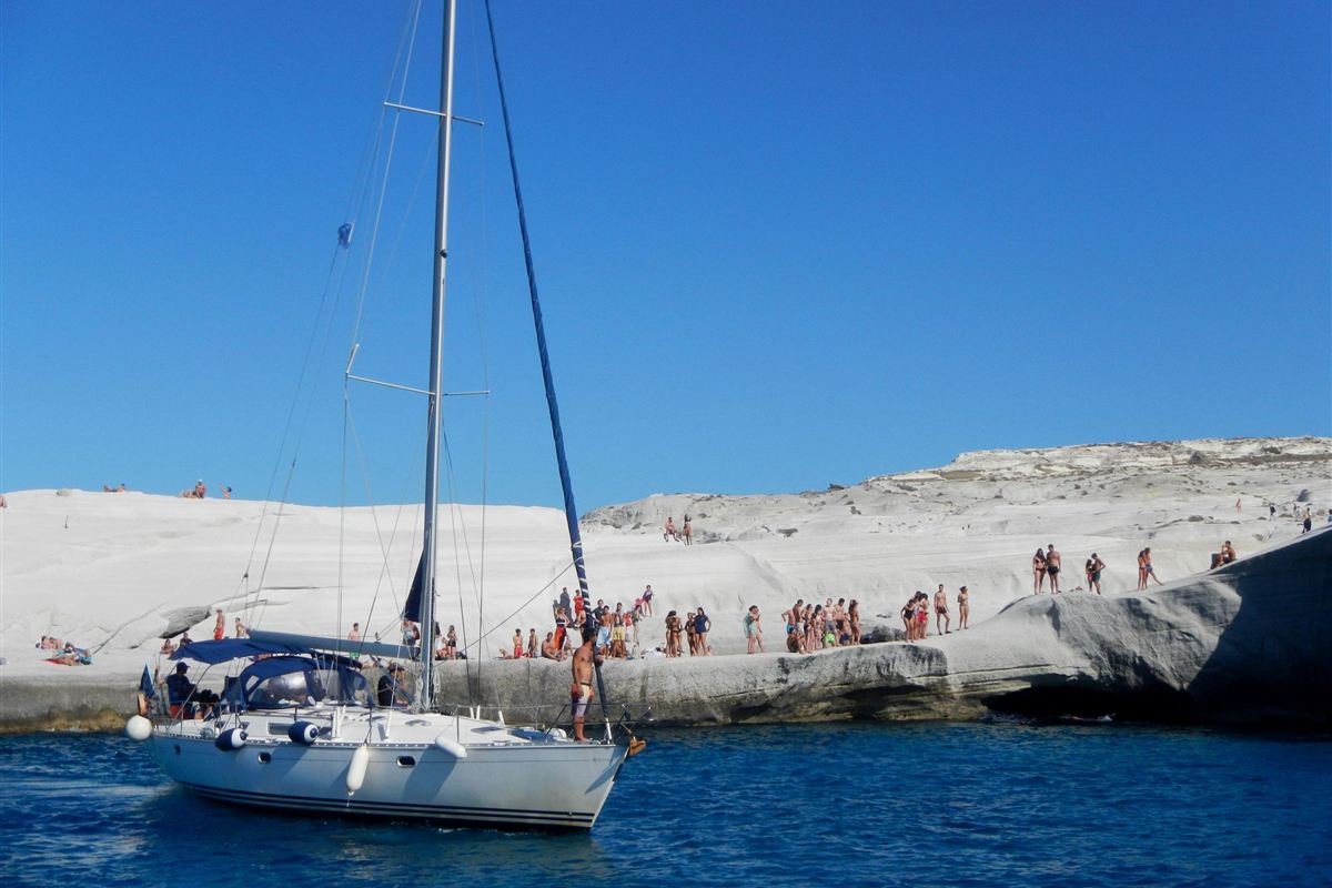 Milos sailing: a white sailboat with a tall mast and blue sail cover cruising close to the smooth white volcanic rock coast of Sarakiniko, with small groups of swimmers on the rocks above deep blue water and a clear blue sky