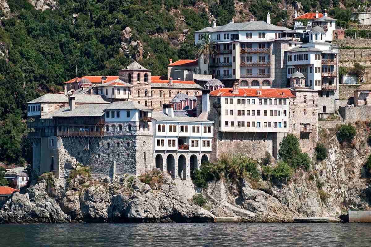 Mount Athos cruise: a grey stone monastery with white upper floors, wooden balconies and orange roofs standing on a rocky cliff above the sea, with a palm tree and dark green forest on the hillside behind