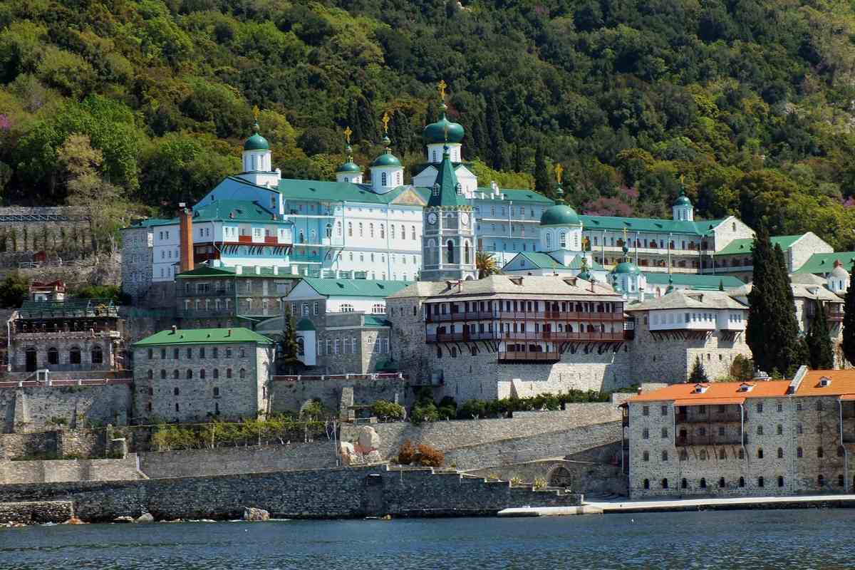 Mount Athos cruise: a monastery with a clock tower, green onion domes and a pale blue church wing above grey stone sea walls, with orange-roofed buildings on the right and a wooded hillside behind blue water