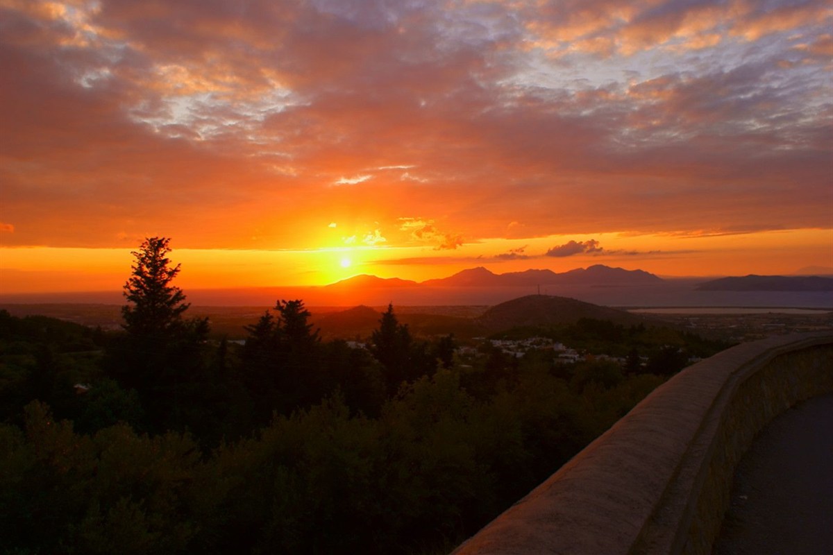 Mount Zia sunset tour: a bright orange sun sinks behind dark mountain silhouettes under a red and orange cloudy sky, seen over dark pine trees, white village houses and the sea, with a curved stone wall beside the road in the foreground