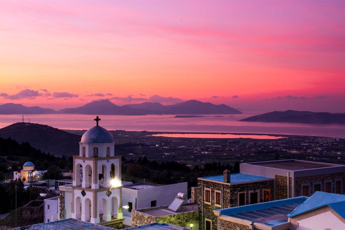 Mount Zia sunset tour: a white and cream village church bell tower with a grey dome and cross rises above flat roofs in Zia while a pink and orange dusk sky glows over dark island silhouettes, the sea and a pink coastal lagoon far below