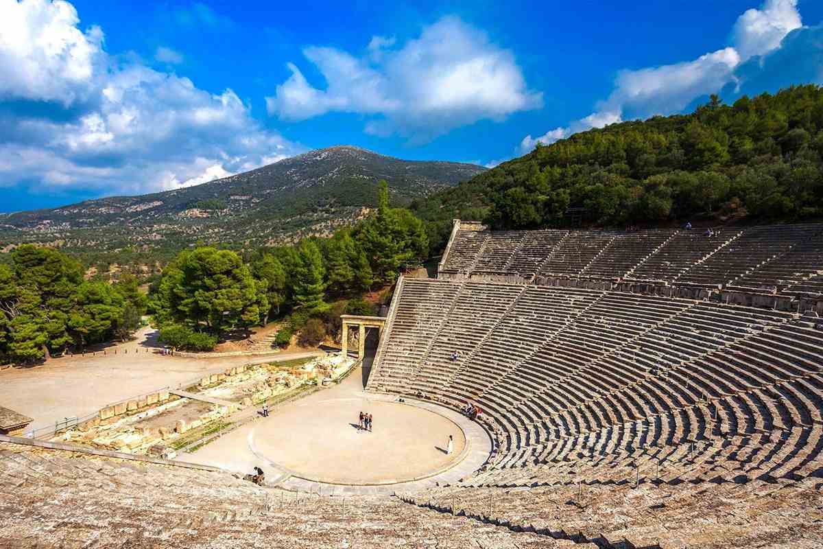 Mycenae Epidaurus Nafplio day trip from Athens: the ancient theatre of Epidaurus seen from the upper tiers, with curved rows of grey stone seats around a round sandy stage, a small stone gateway and pine trees on the left, and a forested mountain under a blue sky with white clouds