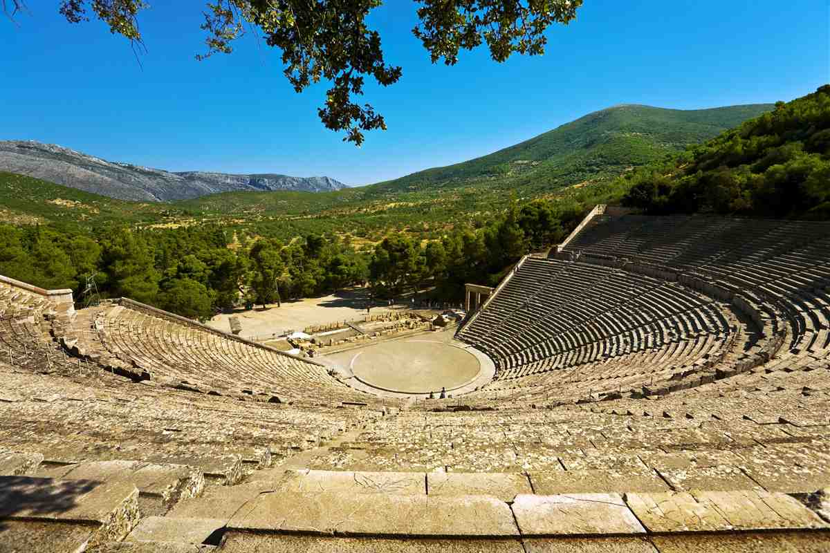 Mycenae Epidaurus tour from Nafplio: the curved rows of grey stone seats of the ancient Theatre of Epidaurus seen from the upper tiers, with a round sandy stage, green pine trees, forested hills and a rocky mountain ridge under a clear blue sky