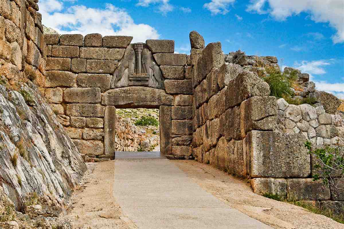 Mycenae Epidaurus tour from Nafplio: the Lion Gate at Mycenae, a doorway of huge cut stone blocks topped by a carved relief of two lions beside a central column, with a pale stone path between rough rock walls under a blue sky with white clouds