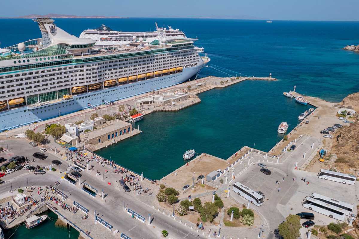 Mykonos shore excursion: a large white and pale blue cruise ship moored at the Mykonos port quay beside a harbour basin of deep green water, with a crowd of passengers on the waterfront road, tour coaches parked on the right and dark blue sea behind
