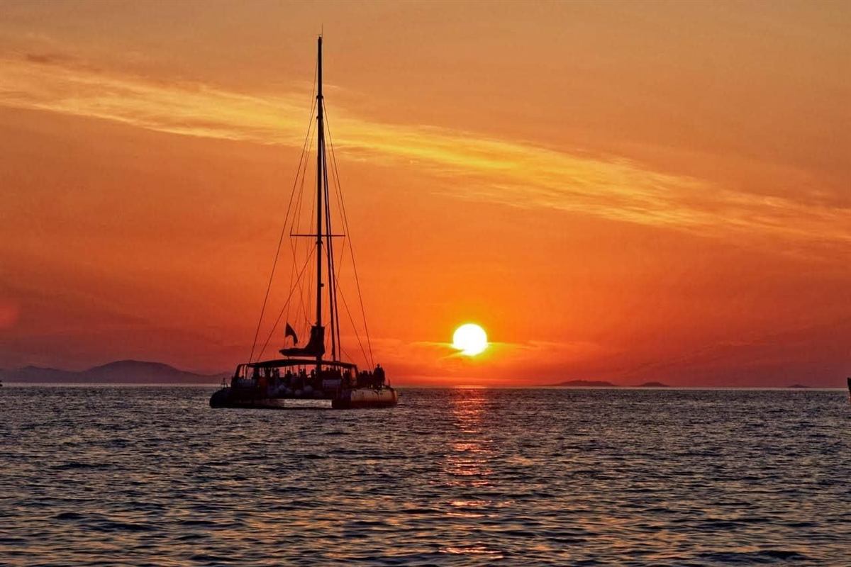 Mykonos sunset cruise: the black silhouette of a two-hulled sailing catamaran with a tall mast and passengers on deck, floating on dark rippled water as a bright white sun sets into a deep orange sky with low islands on the horizon