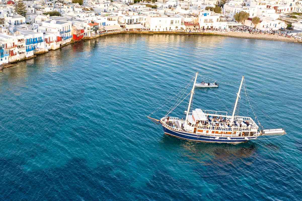 Mykonos sunset cruise: an aerial view of a navy-hulled wooden cruise boat with two white masts and passengers on deck, anchored on deep blue water beside the white waterfront houses and blue balconies of Little Venice in Mykonos Town