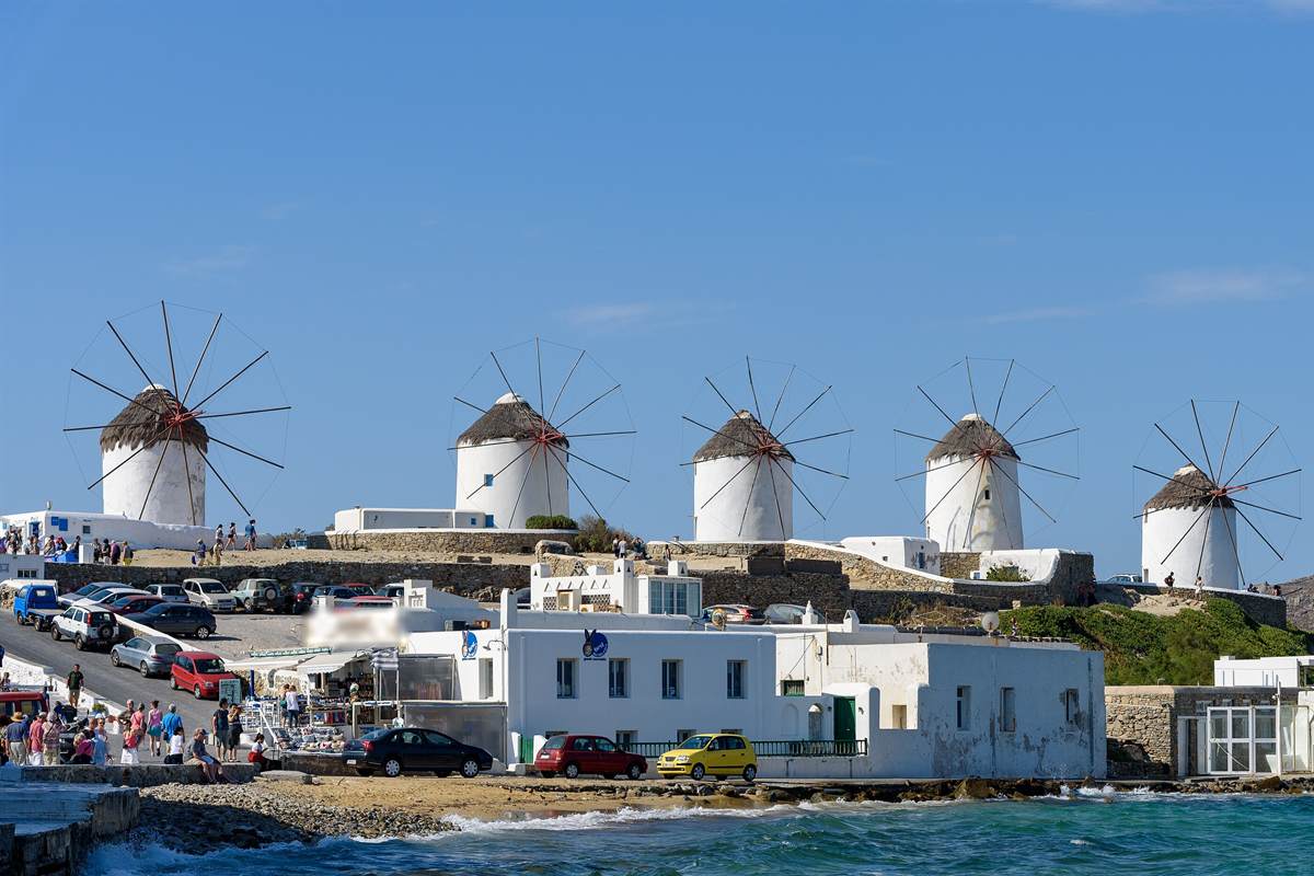 Mykonos wine tasting: five whitewashed windmills with dark thatched roofs and spoked sails on the Kato Mili ridge above a rocky shore, with white buildings below and turquoise sea in the foreground under a clear blue sky