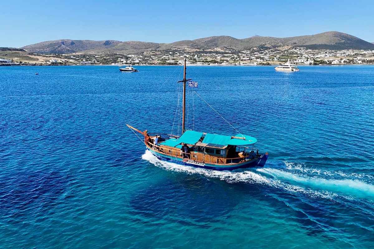 Naoussa bay cruise: a wooden two-masted caique with a turquoise canopy and a Greek flag sailing through deep blue water in Naoussa Bay, with the whitewashed houses and brown hills of the Paros coast behind under a clear blue sky
