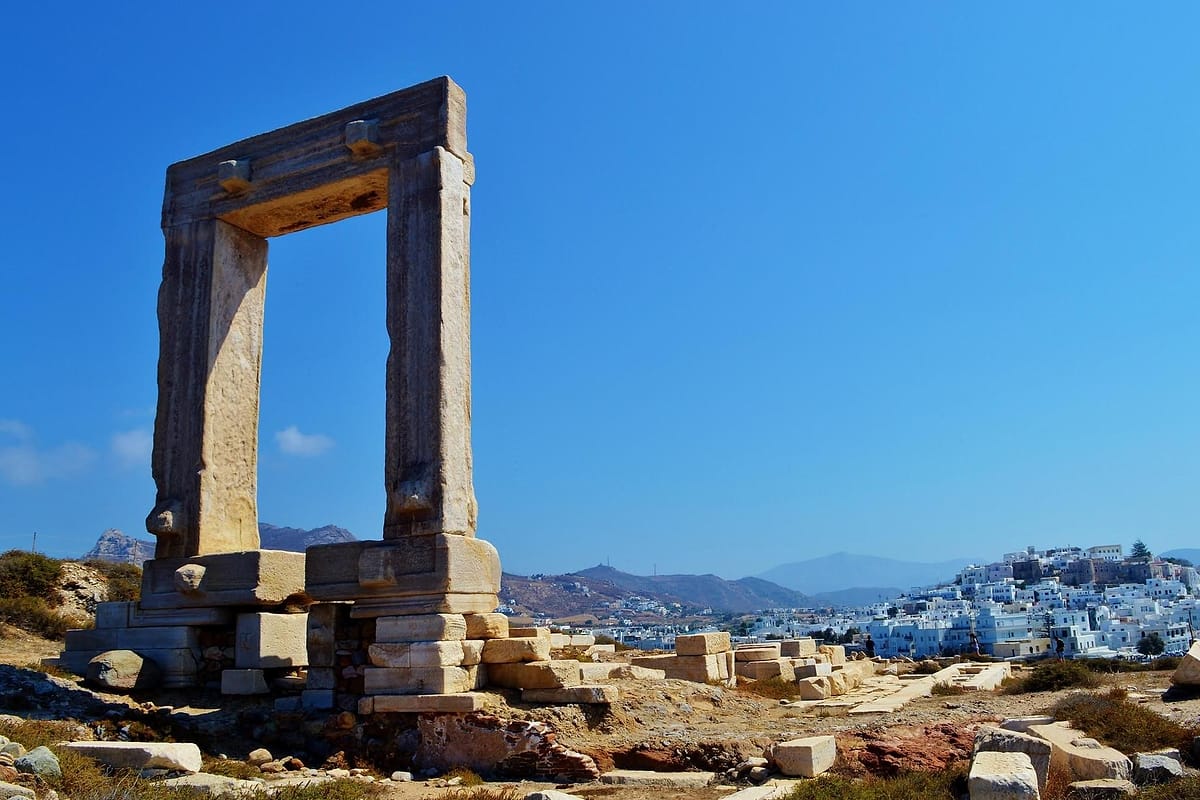 Naxos catamaran cruise: the marble Portara gate on its hill above the white houses of Naxos Town under a bright blue sky