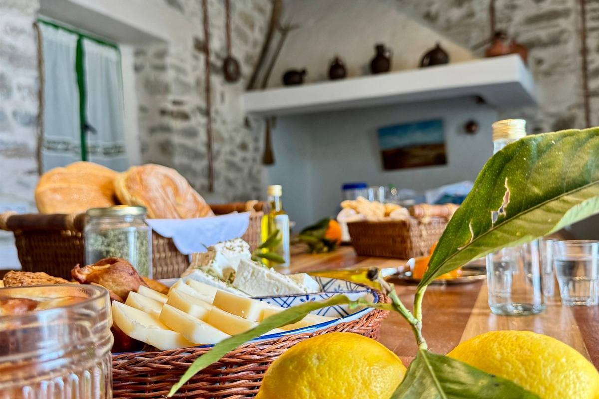 Naxos cooking class: a wooden farm kitchen table in a stone house with a wicker basket of sliced yellow cheese, white feta and crusty bread, a bottle of olive oil, glasses of water and two yellow lemons on a leafy branch