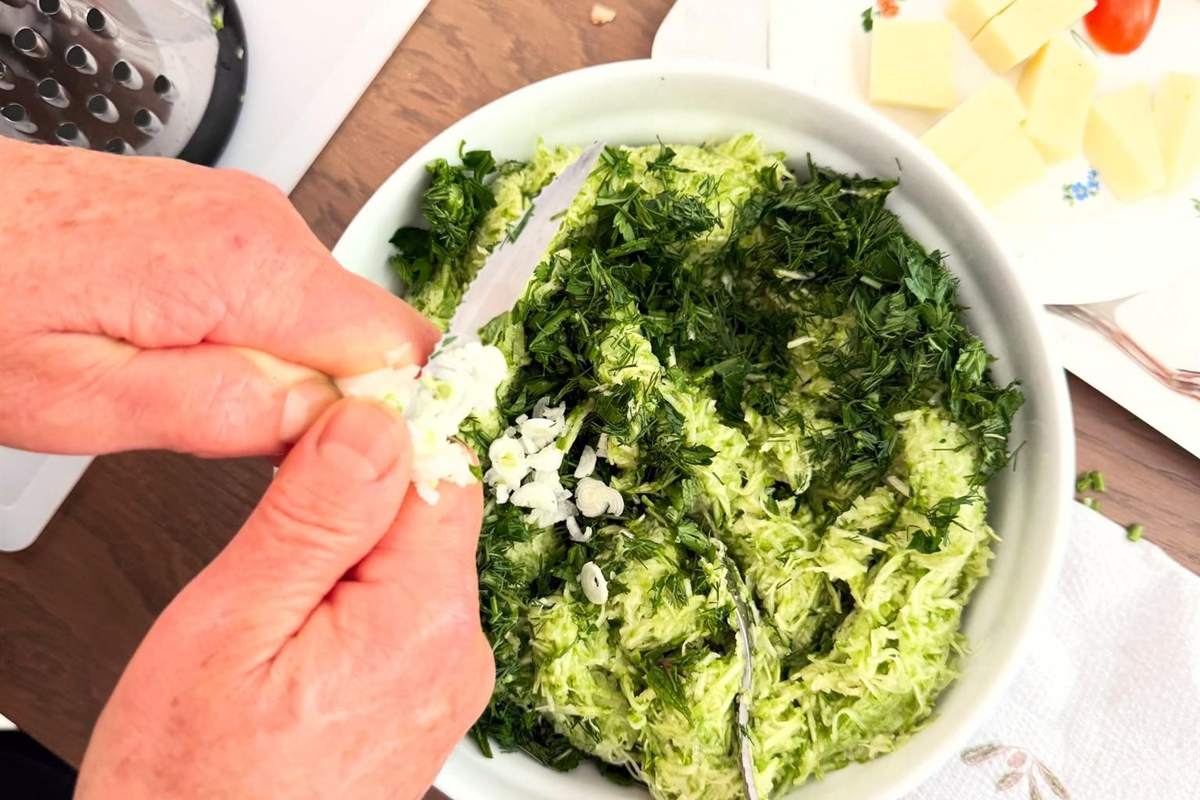 Naxos cooking class: two hands pressing chopped garlic into a white bowl of grated green courgette and fresh dill, with a metal grater and cubes of yellow cheese on a wooden table beside it