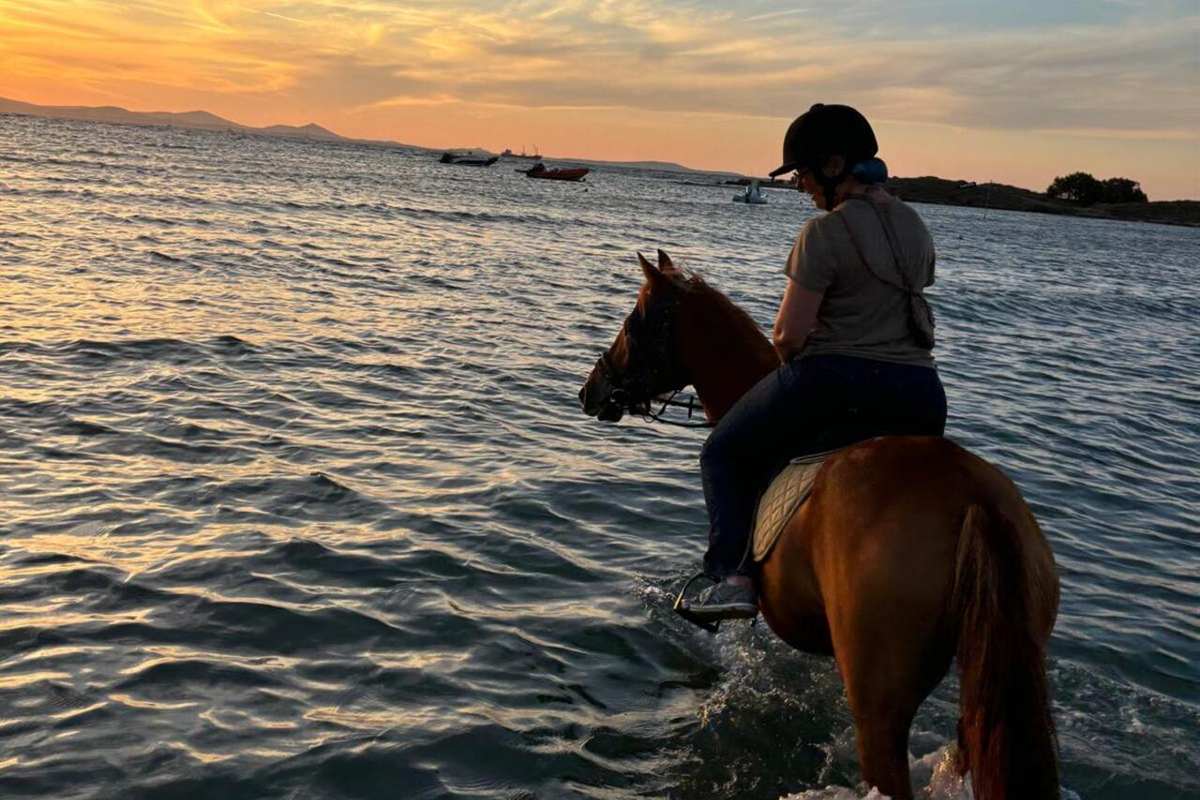 Naxos horse riding: a rider in a dark helmet and grey t-shirt on a chestnut horse walking through rippled shallow sea water under an orange and blue sunset sky, with small boats and a low headland in the distance