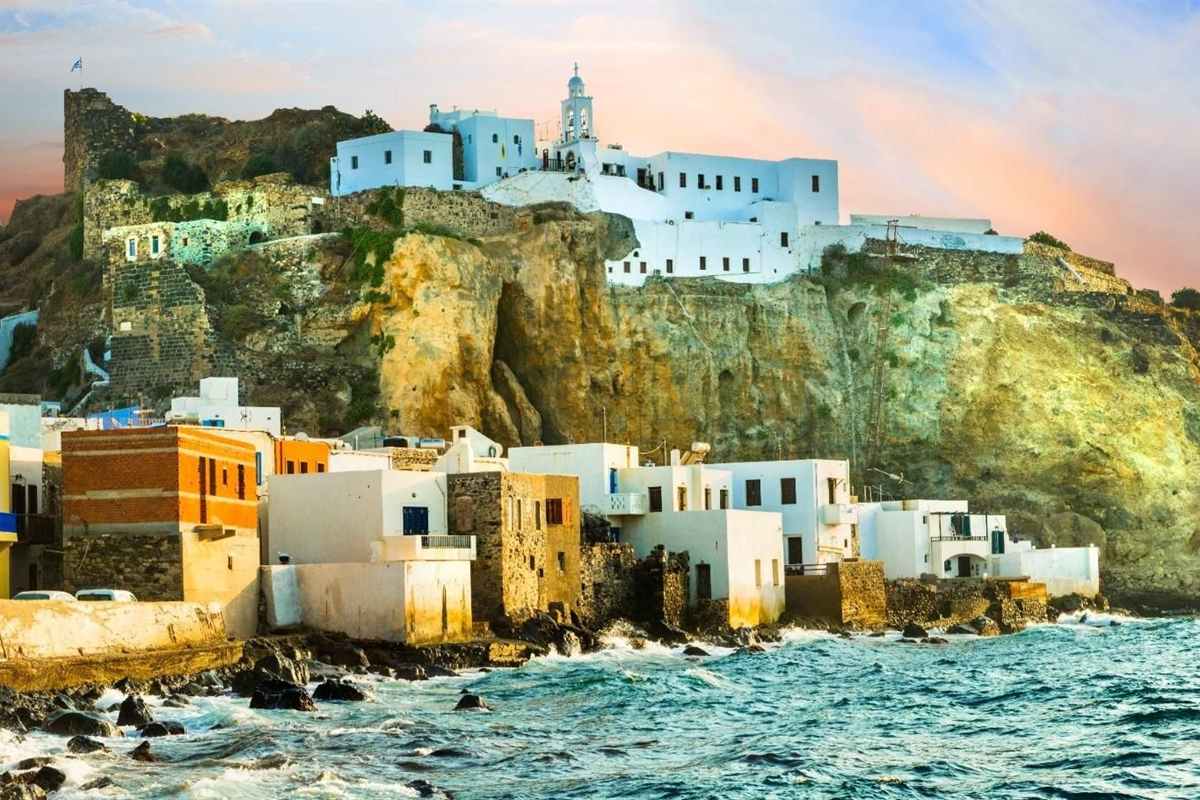 Nisyros day trip from Kos: the whitewashed Panagia Spiliani monastery with its bell tower on a steep golden cliff above Mandraki, a ruined stone castle wall on the left, white and brick-orange houses at the foot of the cliff and waves breaking on dark rocks under a pink and blue sky