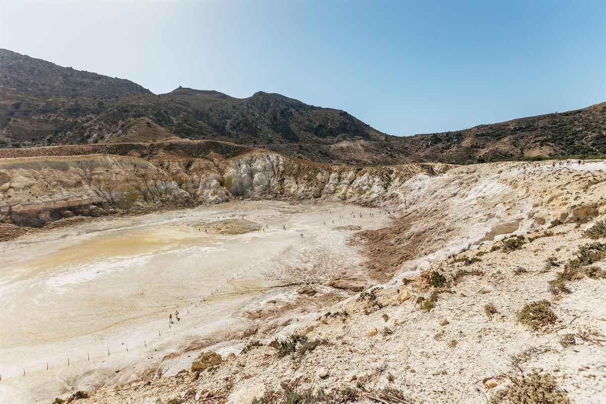 Nisyros day trip from Kos: the wide pale cream and yellow floor of the Stefanos crater seen from the rim, with a fenced path and tiny walkers crossing it, banded white and ochre crater walls behind and dark brown hills under a clear blue sky
