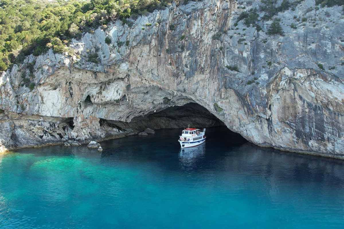 Nydri pig island cruise: a white two-deck passenger boat floating in front of the wide arched mouth of a sea cave in a grey and cream limestone cliff, with green shrubs on the clifftop and deep blue and turquoise water in the foreground