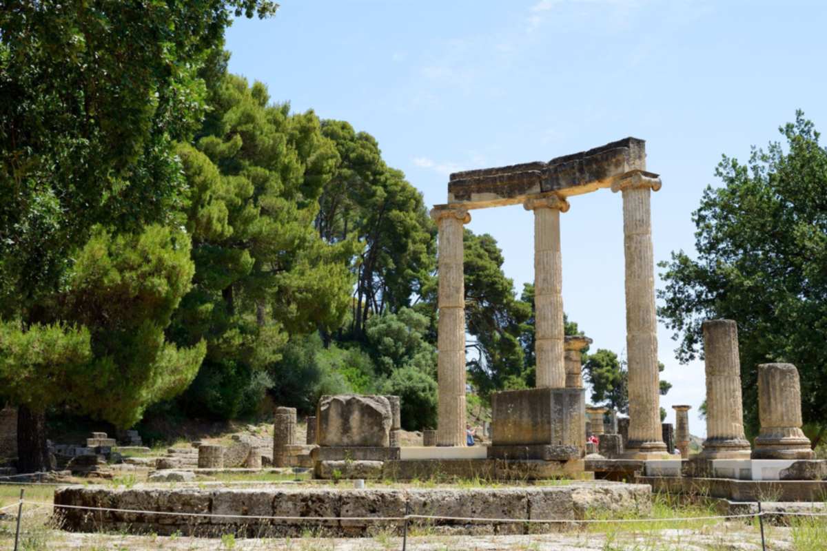 Olympia tickets: the three slender fluted Ionic columns of the round Philippeion at Ancient Olympia carrying a stone beam, with the circular stone base and fallen column drums in front, green pine and oak trees behind and a clear blue sky