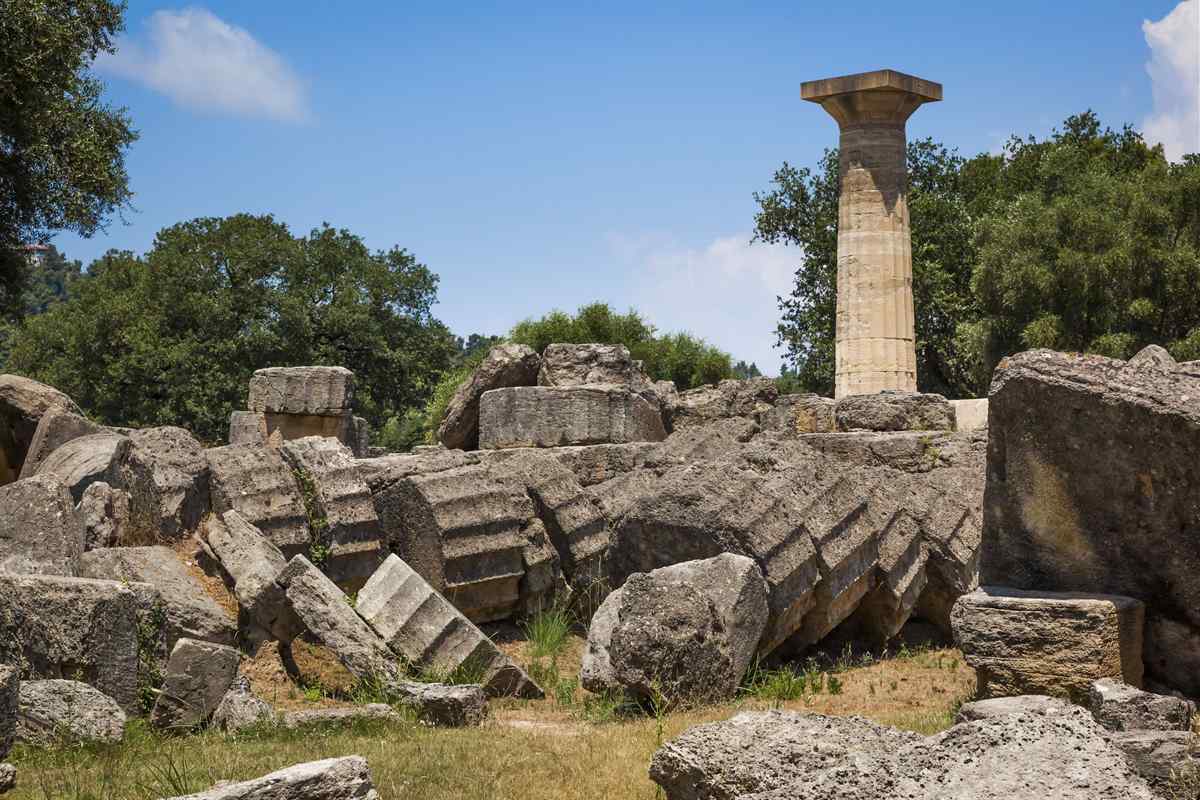 Olympia tickets: the single standing column of the Temple of Zeus at Olympia rising above a field of huge fallen column drums and stone blocks, with green olive trees behind and a blue sky with light clouds
