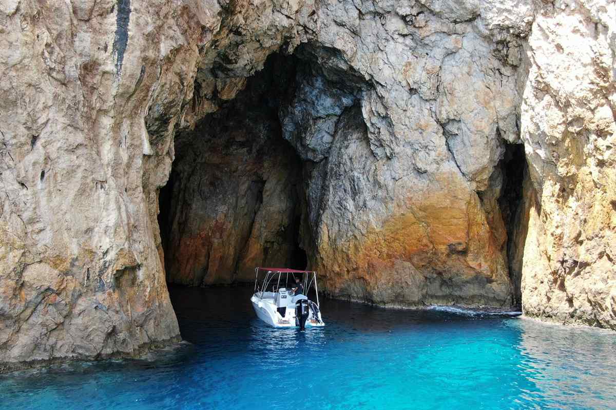 Paleokastritsa boat trips: a small white boat with a red canopy at the mouth of a large sea cave in the layered golden-brown cliffs near Paleokastritsa, with deep blue and turquoise water in front