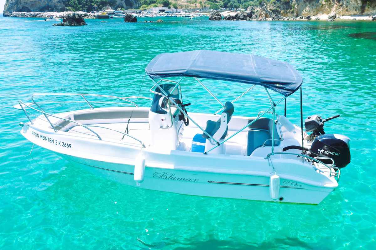 Paleokastritsa boat trips: a small white open boat with a blue canopy, steering console and outboard motor floating on clear turquoise water in a Paleokastritsa bay, with a rocky shoreline and moored boats in the background