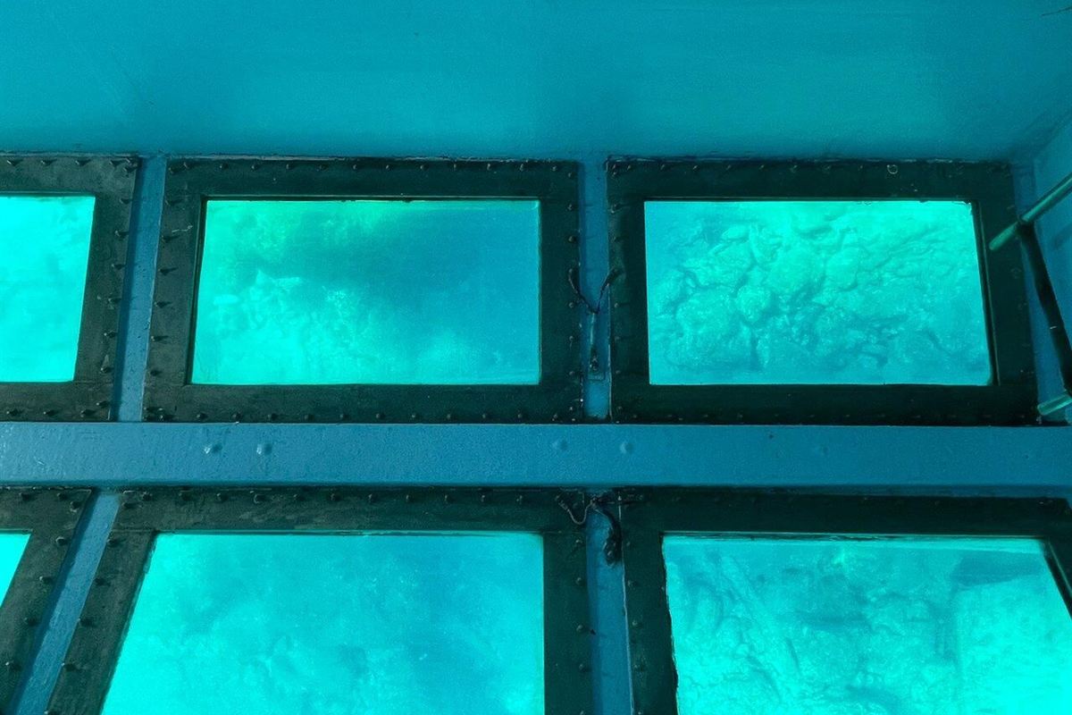 Paphos glass bottom boat: a view inside the hull of two rows of glass panels set in dark riveted frames on a blue steel deck, with turquoise water and a rocky seabed showing through each pane