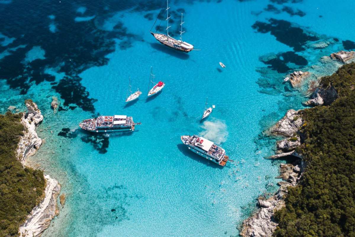 Paxos Antipaxos Blue Caves cruise from Corfu: aerial view of two cruise boats, a gulet and sailboats anchored in a turquoise Antipaxos bay between wooded rocky headlands