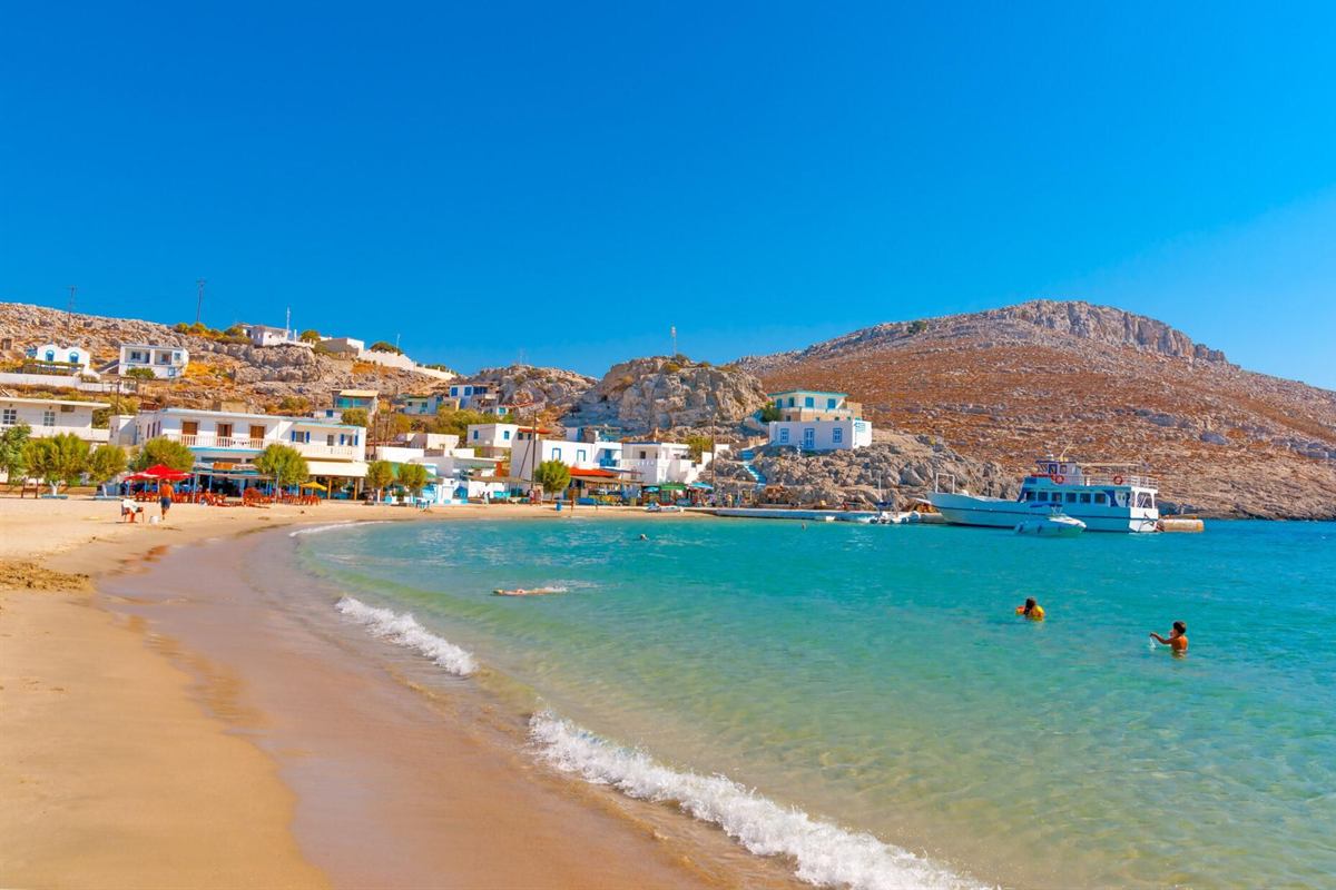 Pserimos day trip from Kos: a curved golden sand beach at Avlakia bay with white and blue houses, tavernas and small green trees along the shore, a white excursion boat at the pier, two swimmers in shallow turquoise water and a rocky brown hill under a deep blue sky