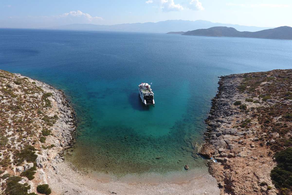 Pserimos day trip from Kos: a white boat with a Greek flag at anchor in clear turquoise water at the mouth of a rocky cove with a pebble beach, dry scrub-covered headlands on both sides and low blue hills across the open sea