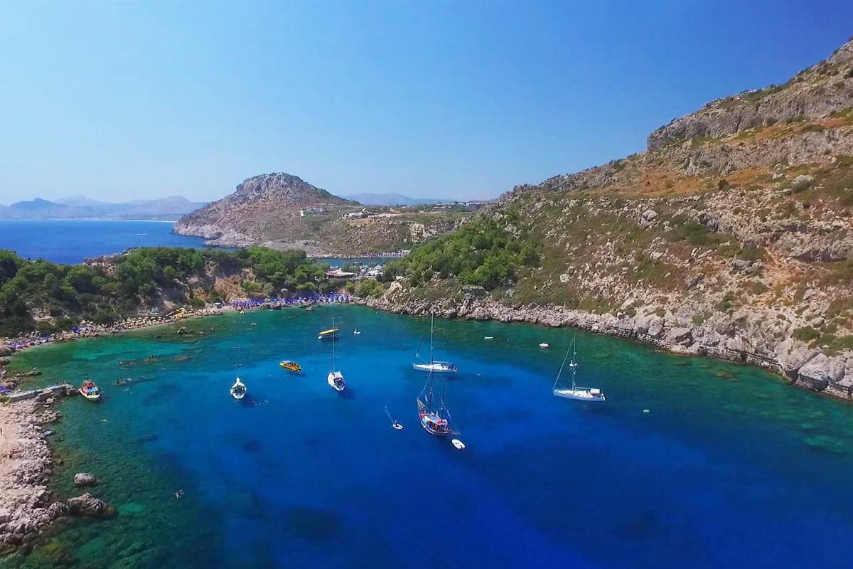 Rhodes catamaran cruise: aerial view of Anthony Quinn Bay with white sailing yachts and a red-hulled wooden cruise boat anchored in deep blue water ringed by turquoise shallows, a pebble beach with blue parasols on the left and rocky pine-covered hills rising on the right