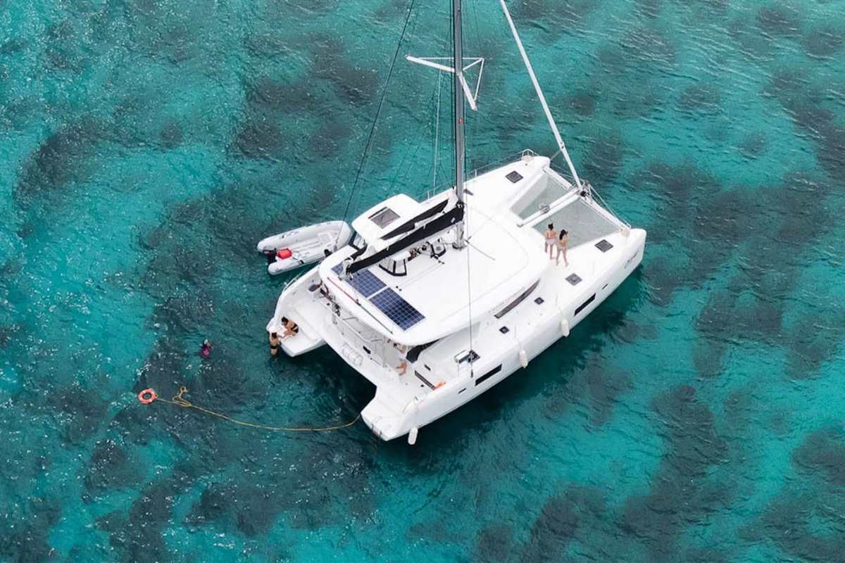 Rhodes catamaran cruise: a white sailing catamaran anchored in clear turquoise water seen from above, with a grey inflatable tender tied alongside, two people standing on the front netting and a swimmer near the stern beside an orange life ring on a long yellow line
