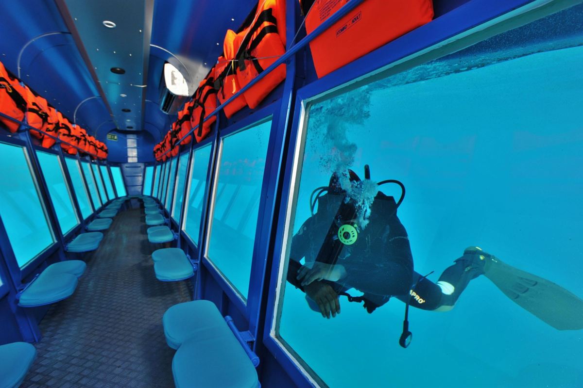 Rhodes submarine cruise: the underwater cabin with blue walls and rows of pale blue seats beside large hull windows, orange life jackets stacked on the racks above, and a scuba diver in a black wetsuit swimming outside one window in clear turquoise water