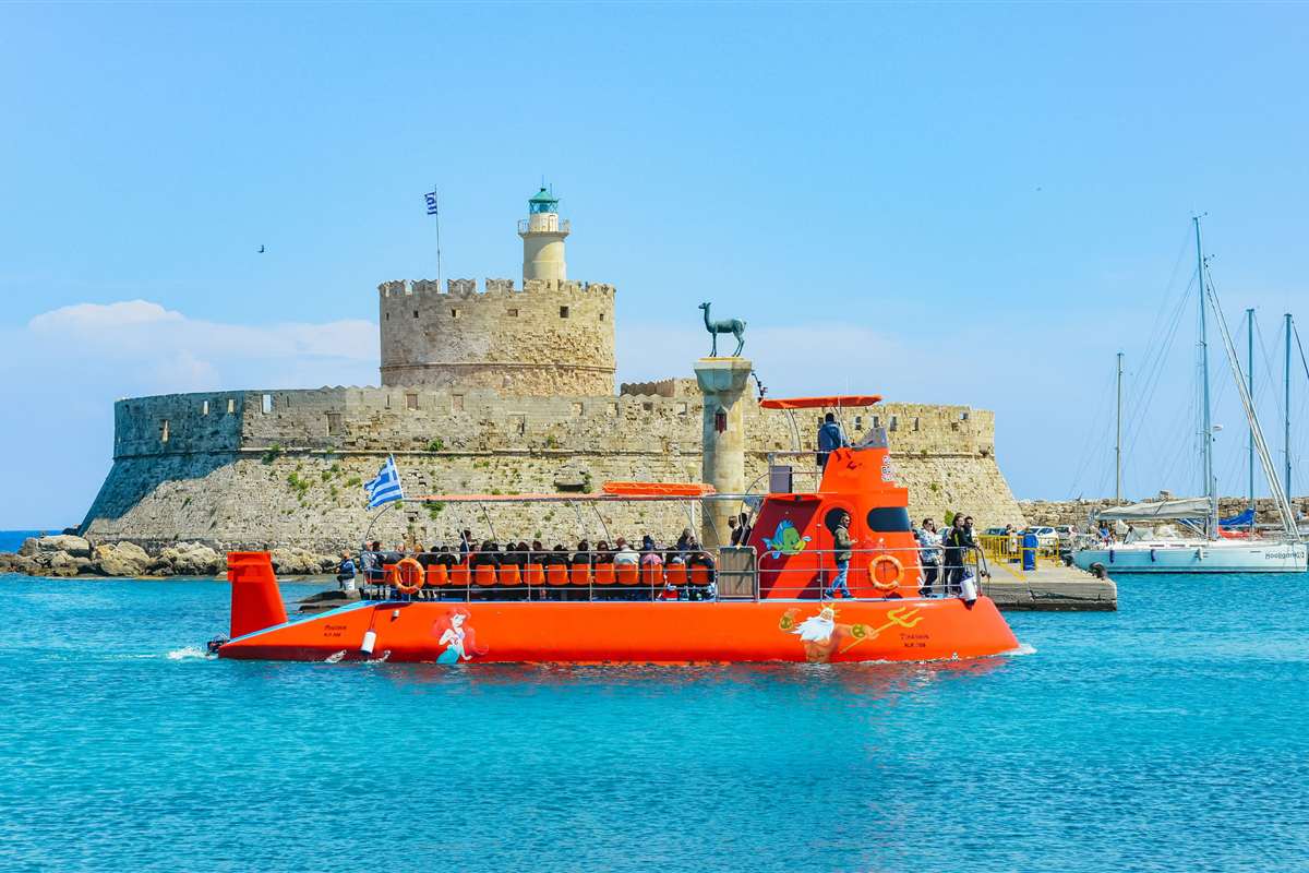Rhodes submarine cruise: a red semi-submarine full of passengers sailing across turquoise water in Mandraki harbour, past the Fort of Saint Nicholas with its round tower and lighthouse and the bronze deer on its column, under a clear blue sky