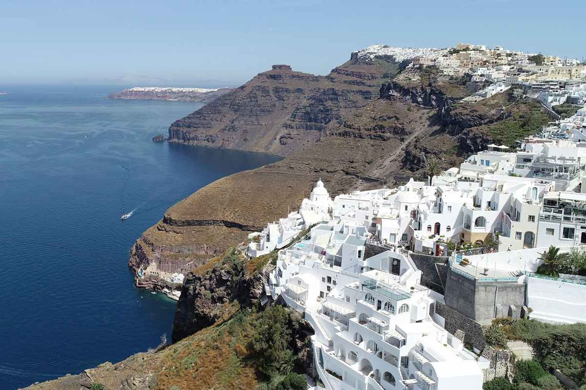 Santorini airport transfer: white cube houses and a small domed chapel of a cliff-top village on the caldera rim, above deep blue water, a brown volcanic headland and a distant white village under a clear blue sky