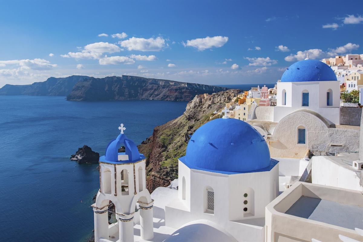Santorini airport transfer: two bright blue church domes and a blue-topped white bell tower of Oia above the deep blue caldera, with pastel houses on the cliff at right and a dark volcanic headland under a blue sky with white clouds