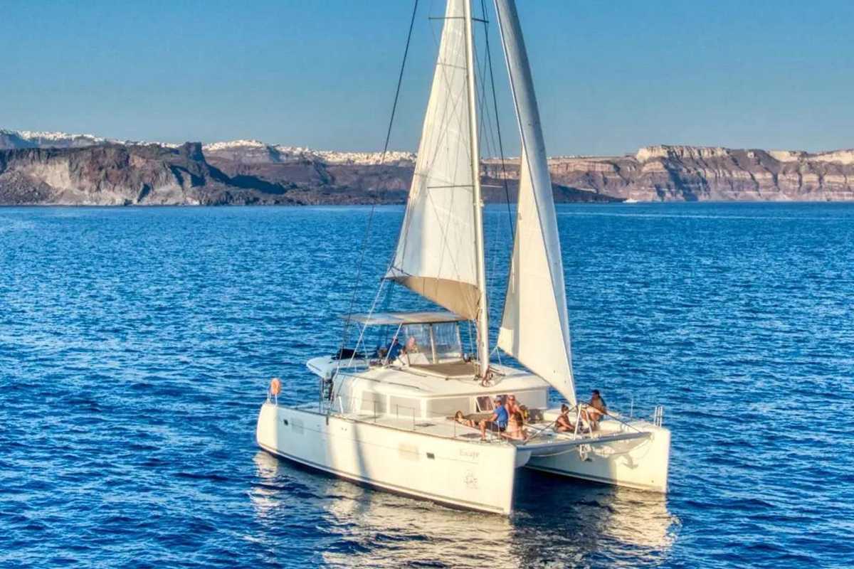 Santorini catamaran cruise with food and open bar: a white catamaran under full sail on the deep blue caldera, with the cliffs and white villages of the caldera rim behind
