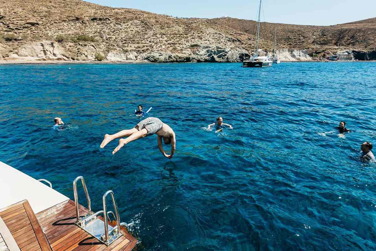 Santorini catamaran cruise with food and open bar: guests swim and one dives into clear blue water off the catamaran stern ladder, with pale volcanic cliffs and a moored catamaran behind