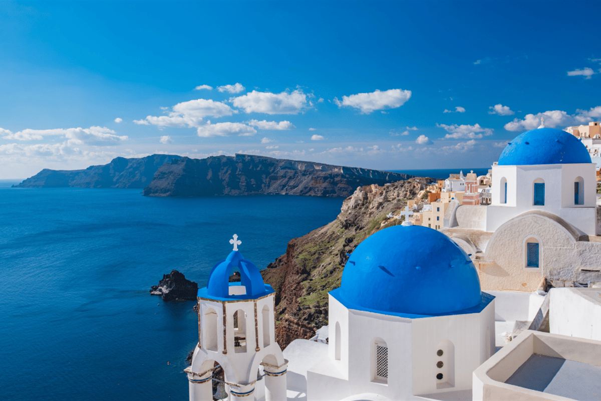 Santorini day trip from Crete: two blue-domed white churches and a blue bell tower at Oia on the caldera cliff, with the deep blue Aegean and the dark caldera rim beyond under a bright sky