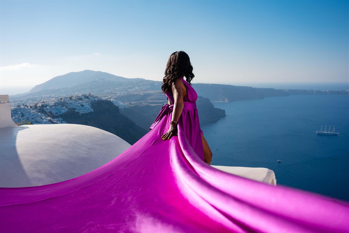 Santorini flying dress photoshoot: a woman seen from behind in a magenta dress, her long train flowing over a white domed roof and terrace edge above the deep blue caldera, with white cliff villages, a dark ridge and a tall sailing ship in the distance