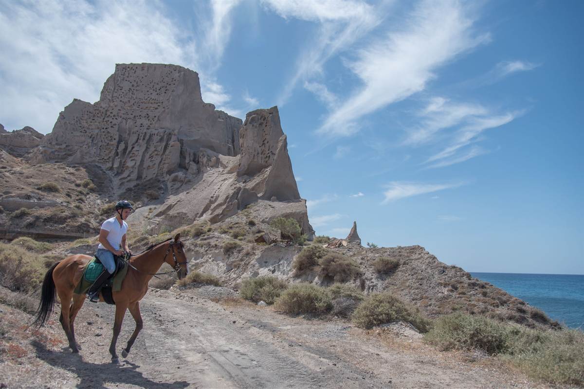 Santorini horse riding: a rider in a dark helmet and white t-shirt on a brown horse with a green saddle pad on a dusty trail, with a tall pale pumice rock full of small holes behind, dry shrubs on the slope and the blue sea at the right under a sky with thin white clouds