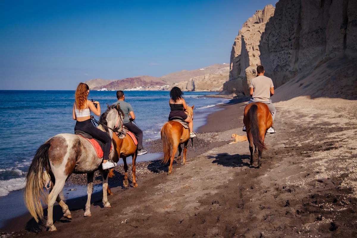 Santorini horse riding: four riders seen from behind on brown and white horses walking along a dark volcanic sand beach beside deep blue sea, with a tall pale pumice cliff on the right, a red-brown headland in the distance and a dog resting on the sand under a clear blue sky