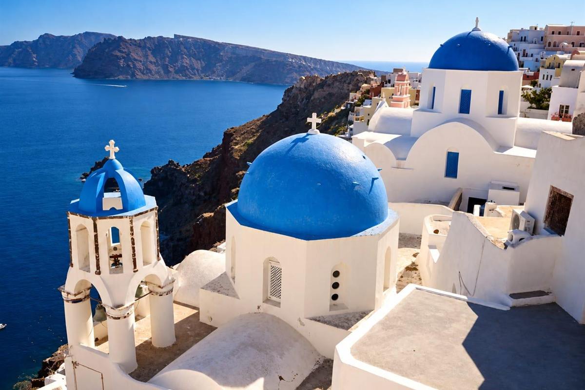 Santorini private tour: two blue-domed whitewashed churches and a white bell tower with a blue cap in Oia, standing above the deep blue caldera water with dark volcanic cliffs across the bay under a clear blue sky