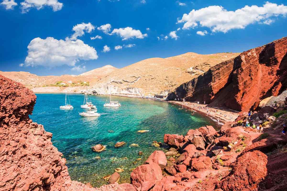 Santorini private tour: a sheltered cove of turquoise water with white sailboats at anchor, framed by red volcanic cliffs and rust-coloured boulders at Red Beach under a bright blue sky with white clouds